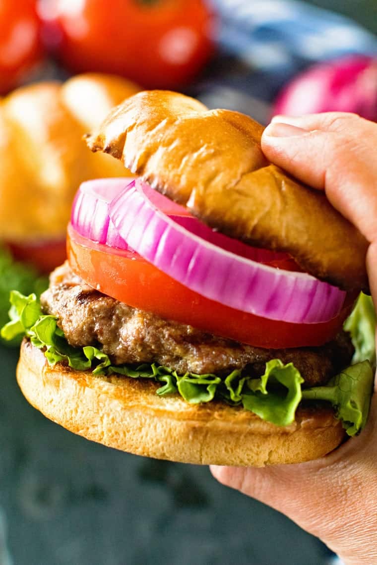 Hand holding a hamburger in a bun toppedw ith onion, tomato and lettuce.