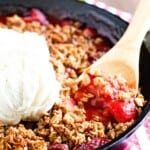 A black cast iron skillet filled with a hot bubbly strawberry crip. A wooden spoon is serving up a spoonful topped with vanilla ice cream.