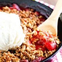 A black cast iron skillet filled with a hot bubbly strawberry crips. A wooden spoon is serving up a spoonful topped with vanilla ice cream.