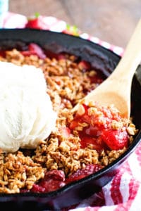 A black cast iron skillet filled with a hot bubbly strawberry crips. A wooden spoon is serving up a spoonful topped with vanilla ice cream.