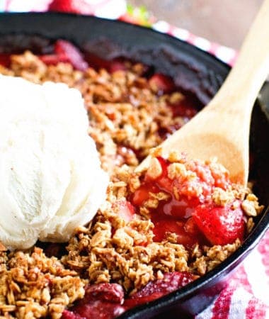 A black cast iron skillet filled with a hot bubbly strawberry crips. A wooden spoon is serving up a spoonful topped with vanilla ice cream.