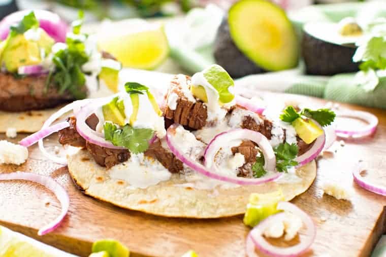 Flank steak tacos on a wooden curling board topping with cilantro, red onion and avocado.