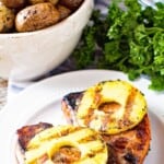 Grilled pineapple pork chops on a white plate with a bowl of potatoes.