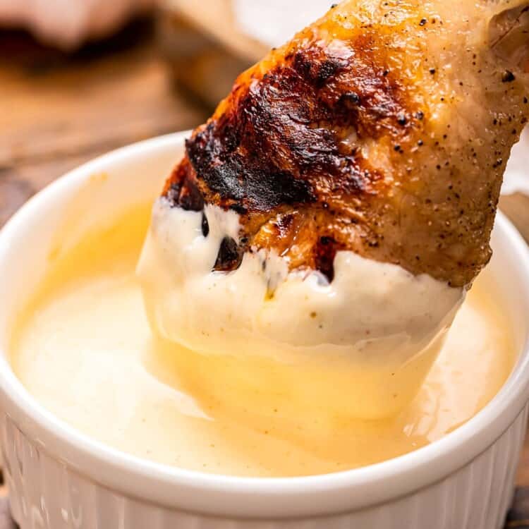 Chicken leg being dunked into Alabama White BBQ Sauce in a white dish