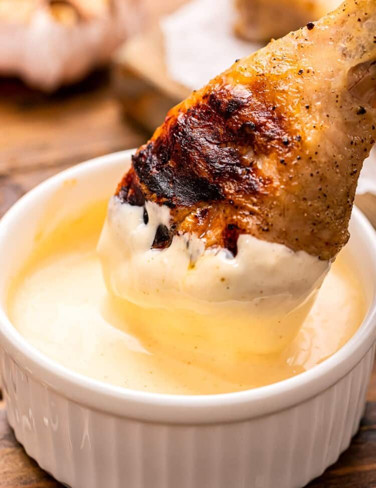 Chicken leg being dunked into Alabama White BBQ Sauce in a white dish