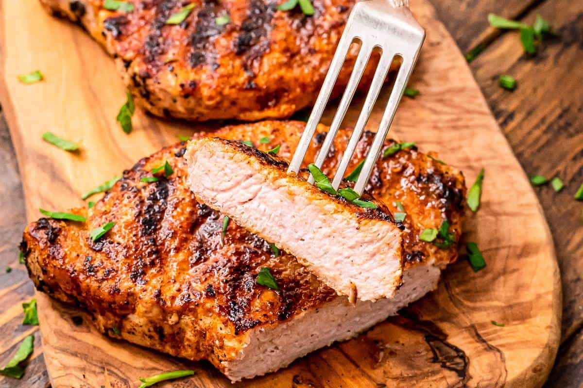 Slice of Pork Chop on fork with the rest of the pork chop in background on wood cutting board