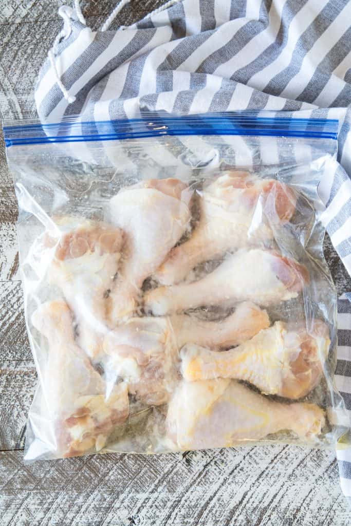 Overhead image of a ziplock back with raw chicken legs in it on a white washed background and blue striped napkin in background
