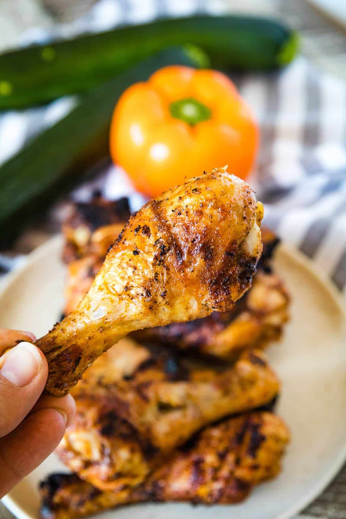 Fingers holding grilled chicken leg with plate of chicken legs in background along with orange pepper and zucchinis