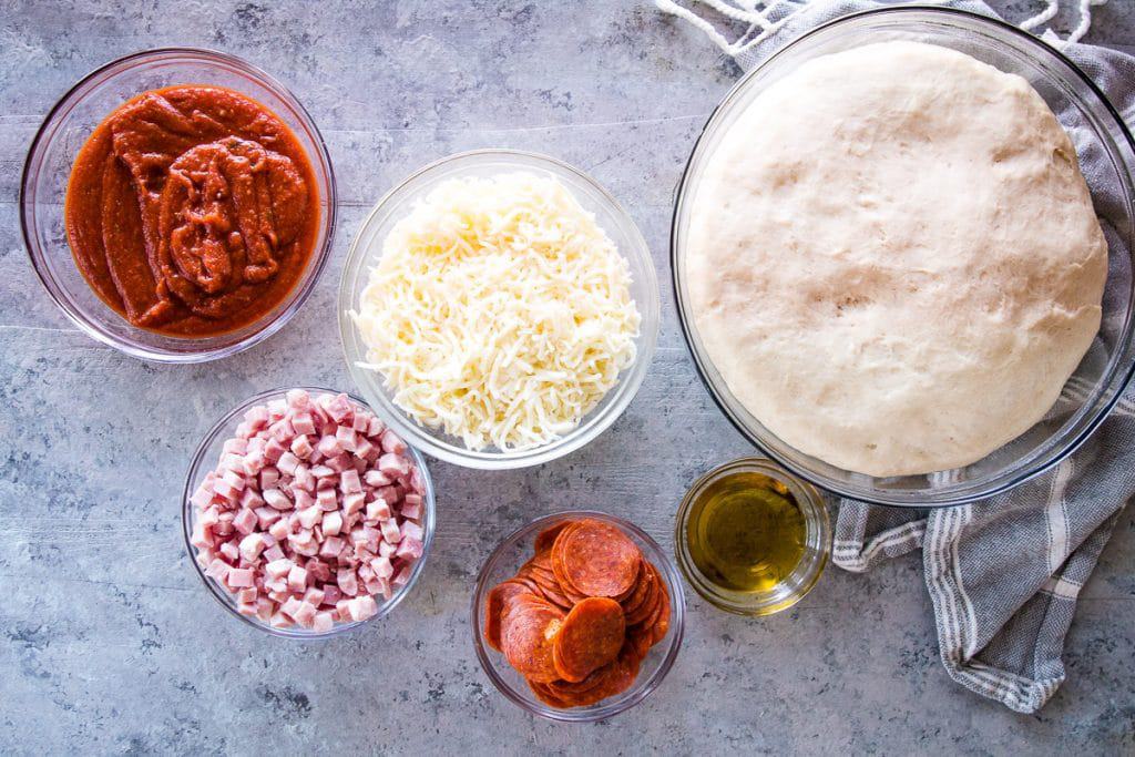 Overhead image showing ingredients in glass bowls to make grilled pizza. Including pizza sauce, cheese, pepperoni, diced ham, olive oil.