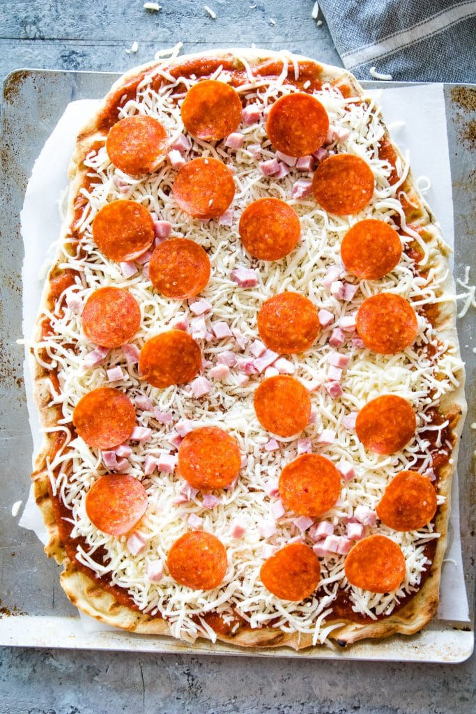 Pizza crust on sheet pan topped with sauce, shredded mozzarella cheese and pepperoni before cooking.