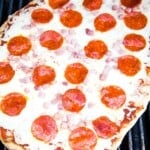 Picture of a pizza on grill grates cooking topped with cheese, diced ham and pepperoni.