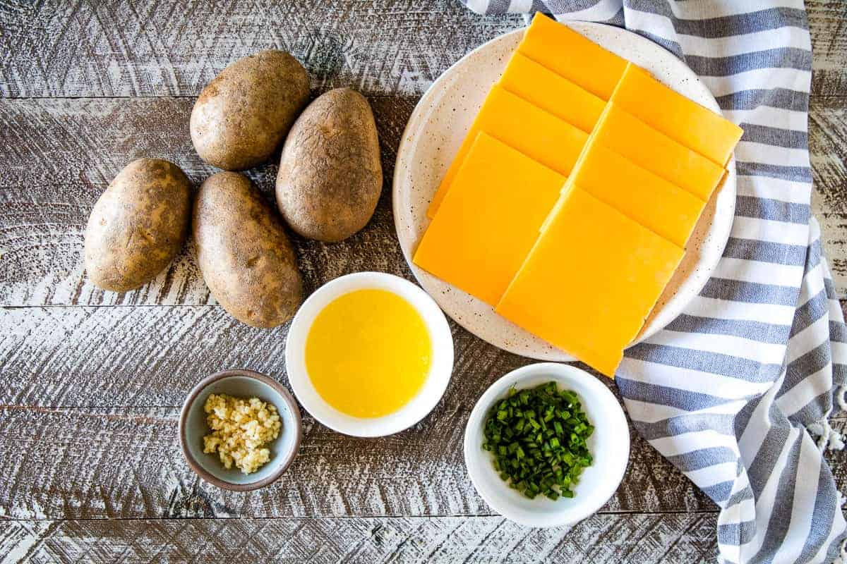 Overhead image of potatoes, slices of cheese on plate, melted butter in bowl, minced garlic in bowl and chopped chives in a bowl.
