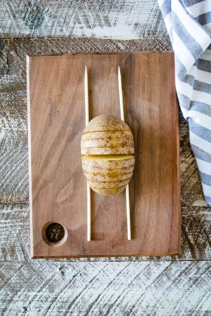 Russet potato sitting on a wood cutting board with chopsticks on each side with slits cut into it.