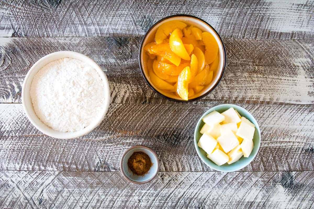 Overhead image of a bowl of dry yellow cake mix, bowl of sliced canned peaches, cinnamon and sliced slabs of butter.