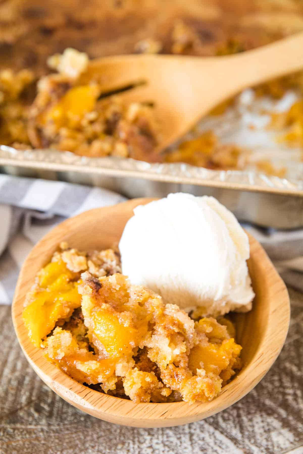 Brown wodden bowl with peach dump cake and a scoop of vanilla ice cream. A pan of more cake behind that.
