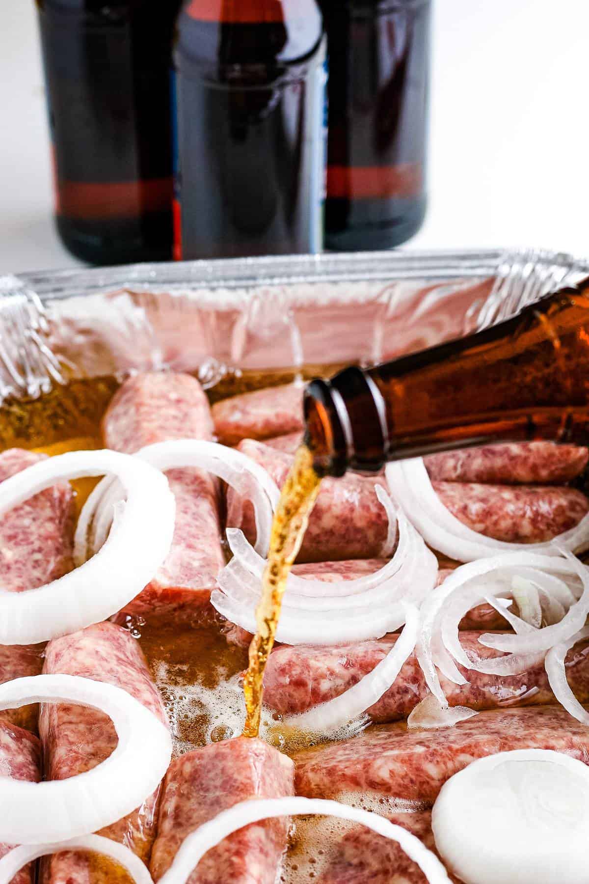 Amber colored bottle of beer being poured into disposable pan with sliced onions and brats.