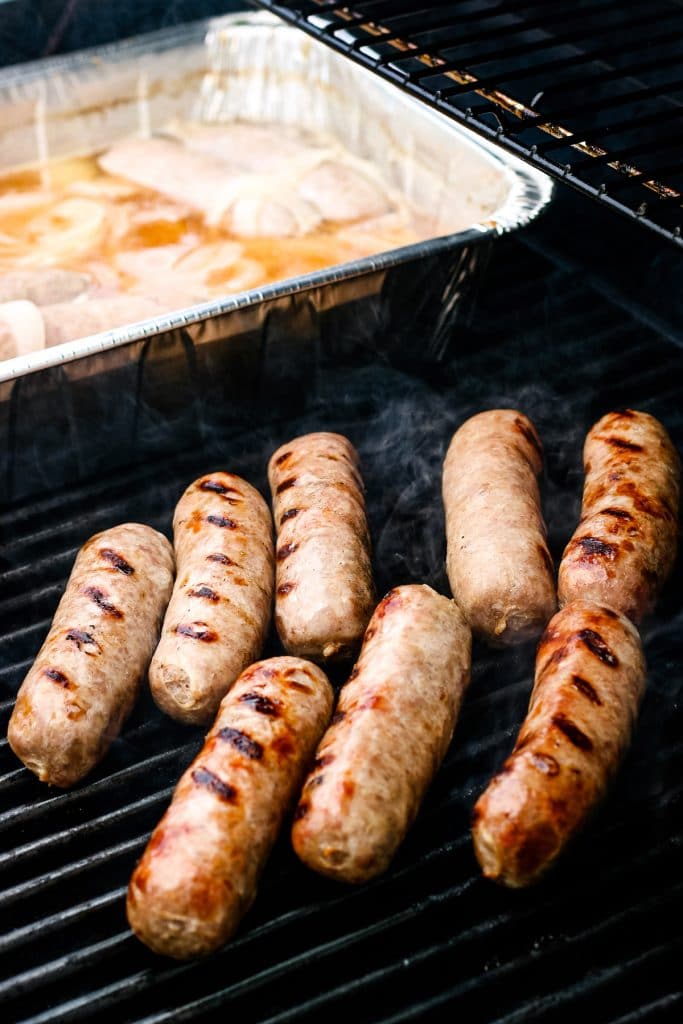 Brats being grilled on grate with a disposable pan of beer and onions in background.
