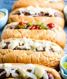 Close up image of five beer brats on buns iwth toppings like mustard, onions, ketuchup and more.