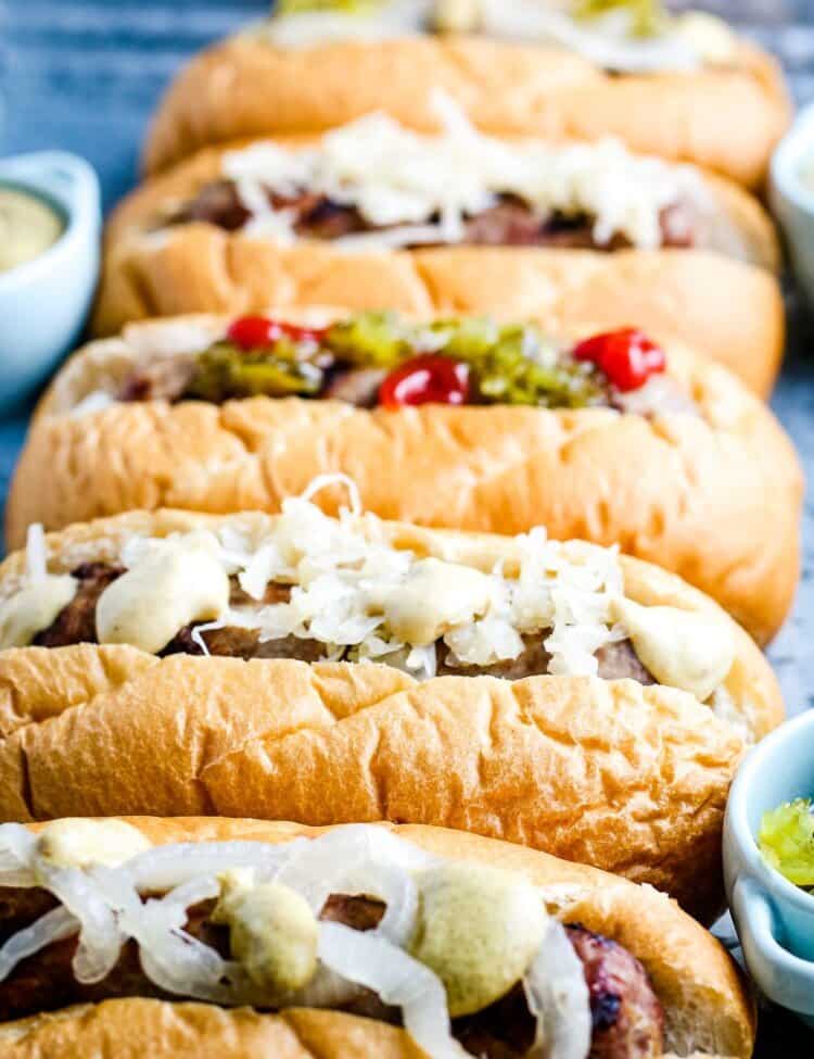 Close up image of five beer brats on buns iwth toppings like mustard, onions, ketuchup and more.