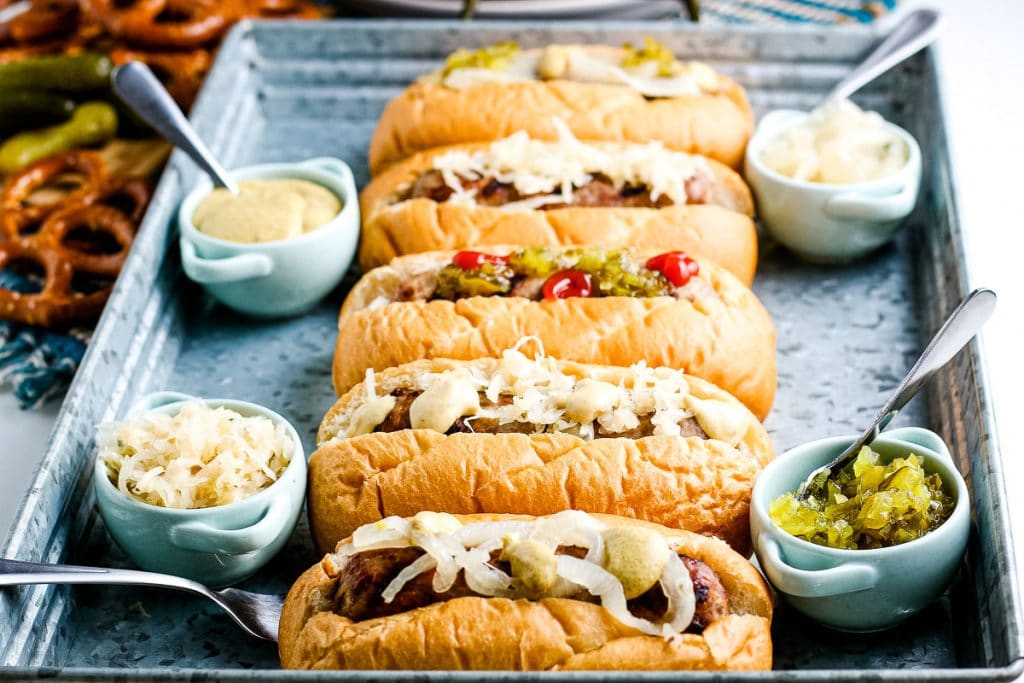 Four brats on buns with varied toppings with small bowls of condiments next to them.