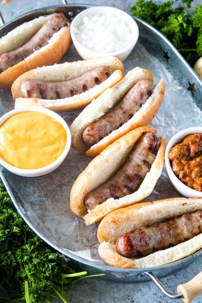 Brats in buns on silver serving tray with toppings next to them.