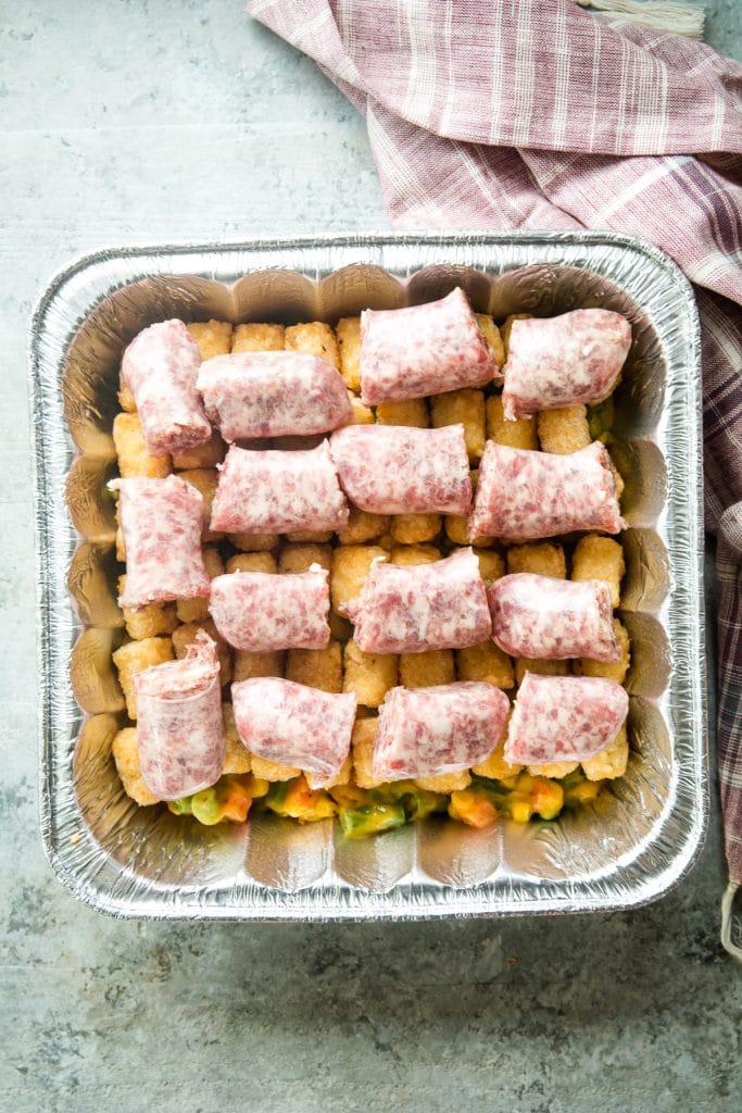 Square foil pan with cheese mixed vegetables in the bottom and topped with frozen tater tots and brat slices.