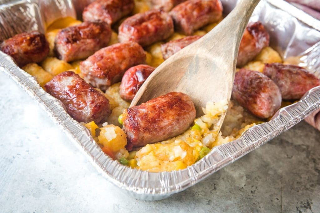 Wooden spoon scooping brat tater tot casserole out of foil pan