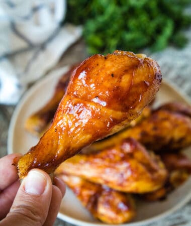 Hand holding Smoked Chicken Legs above plate of chicken legs