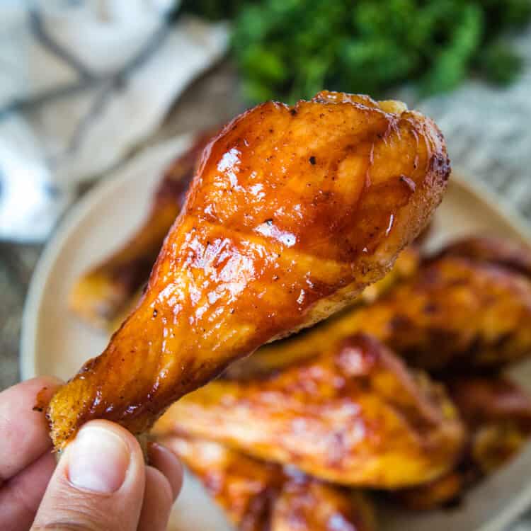 Hand holding Smoked Chicken Legs above plate of chicken legs