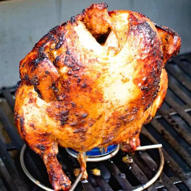 Grilled whole chicken stuffed with beer can sits upright on a grill.