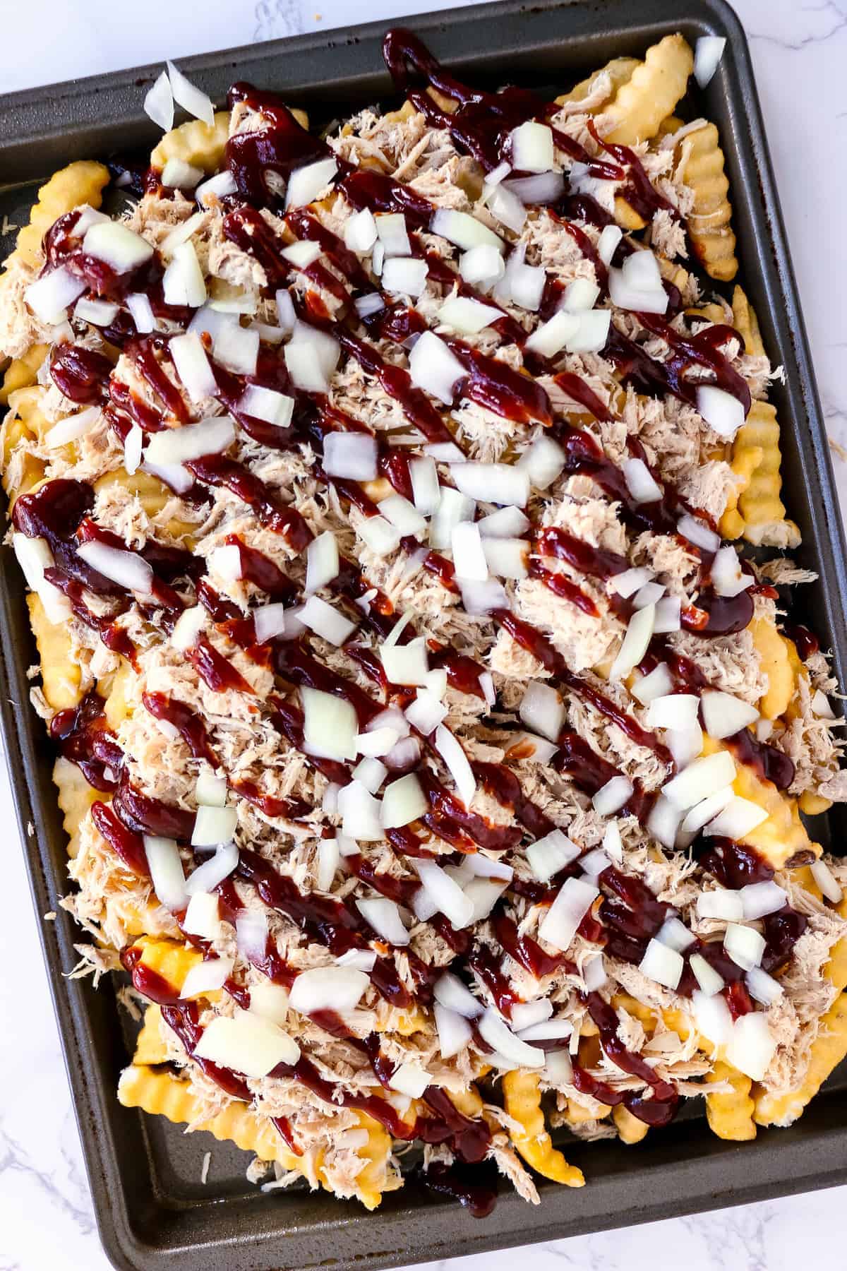 Sheet pan with frozen french fries, pulled pork, BBQ sauce and onions on it