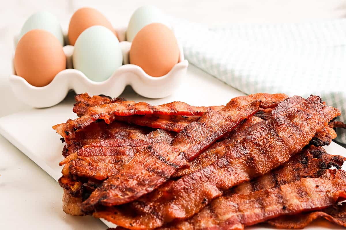 Grilled bacon stacked on plate with eggs in background