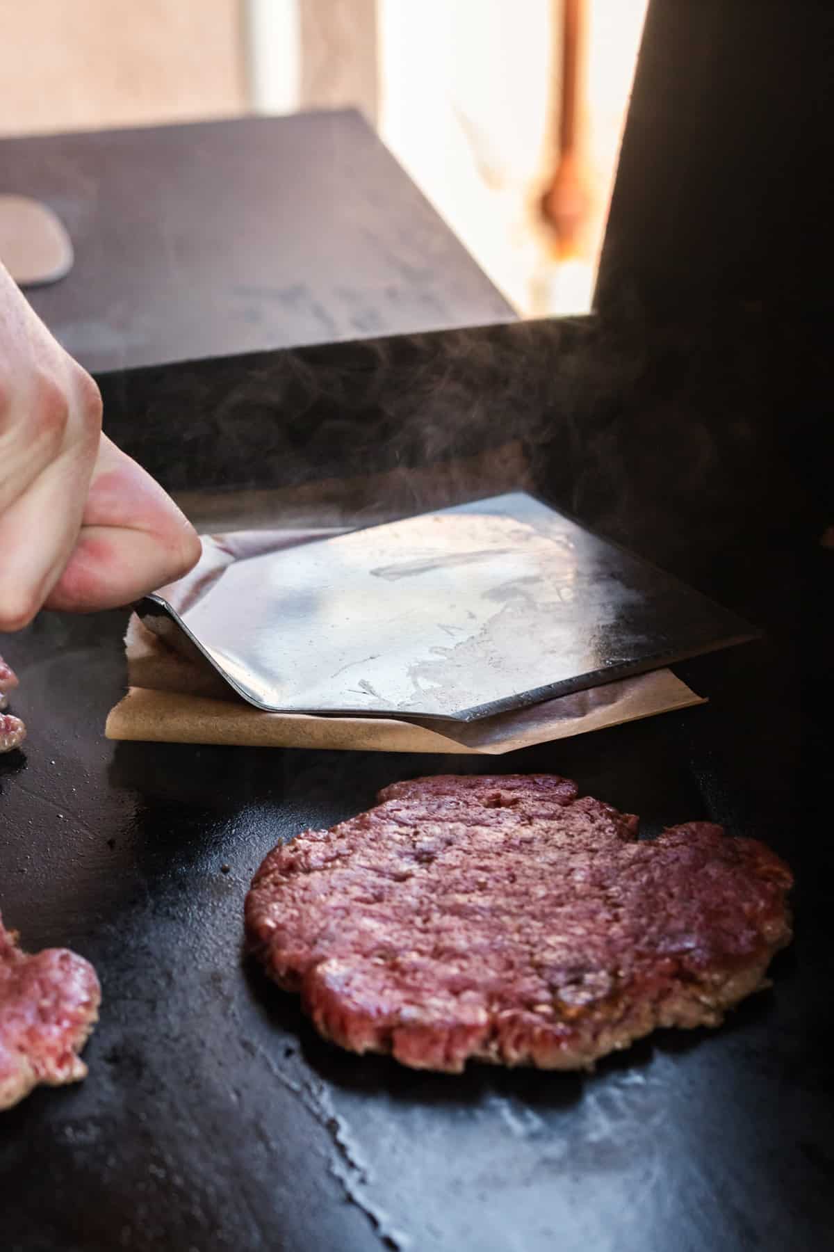 Placing patties on Blackstone griddle and pressing down on parchment paper with spatula for Blackstone Patty Melts