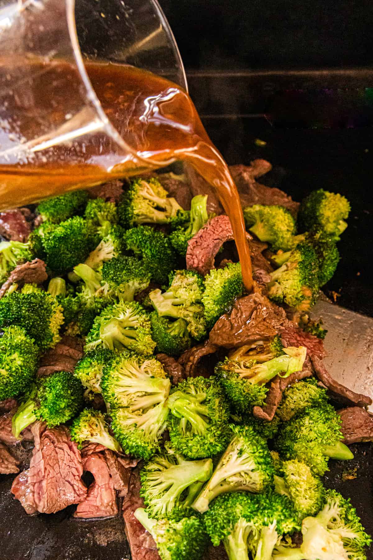 Pouring Blackstone Beef & Broccoli sauce over cooked ingredients on griddle