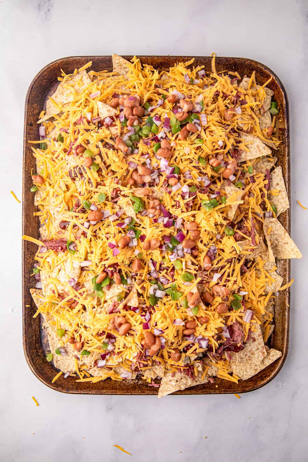 Loading the Smoked Brisket Nachos toppings with shredded cheddar cheese on baking sheet