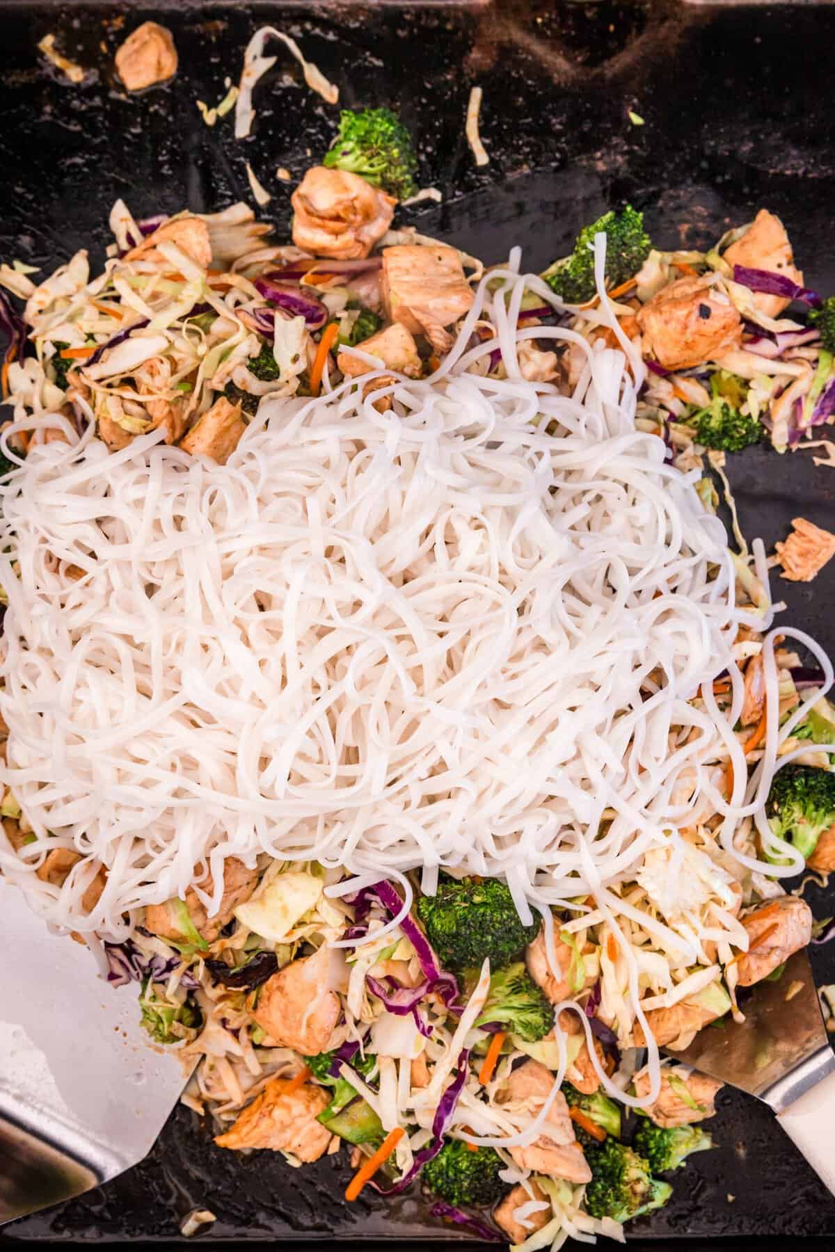 Adding cooked rice noodles to ingredients on the griddle for Blackstone Chicken Stir Fry recipe