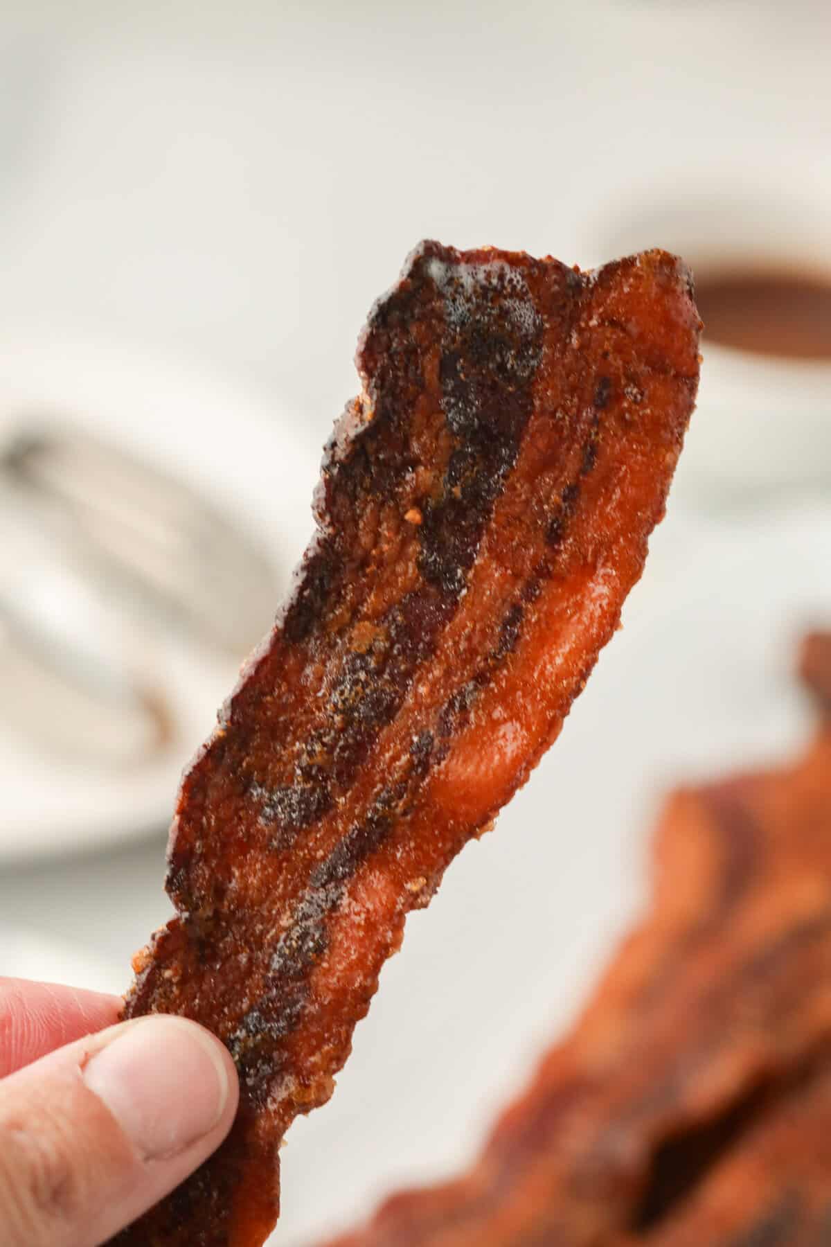 Picture of hand holding up a single piece of completed Smoked Candied Bacon.