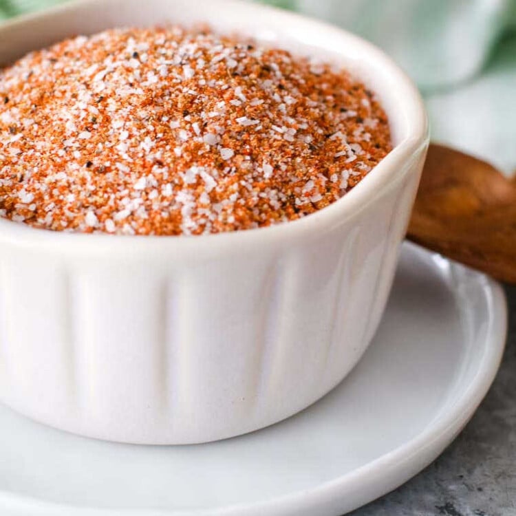 A small white ramekin filled with a homemade dry rub mixture, consisting of coarse salt, black pepper, and various spices, giving it a reddish-brown color. The ramekin sits on a white plate with a wooden spoon nearby, against a blurred background.