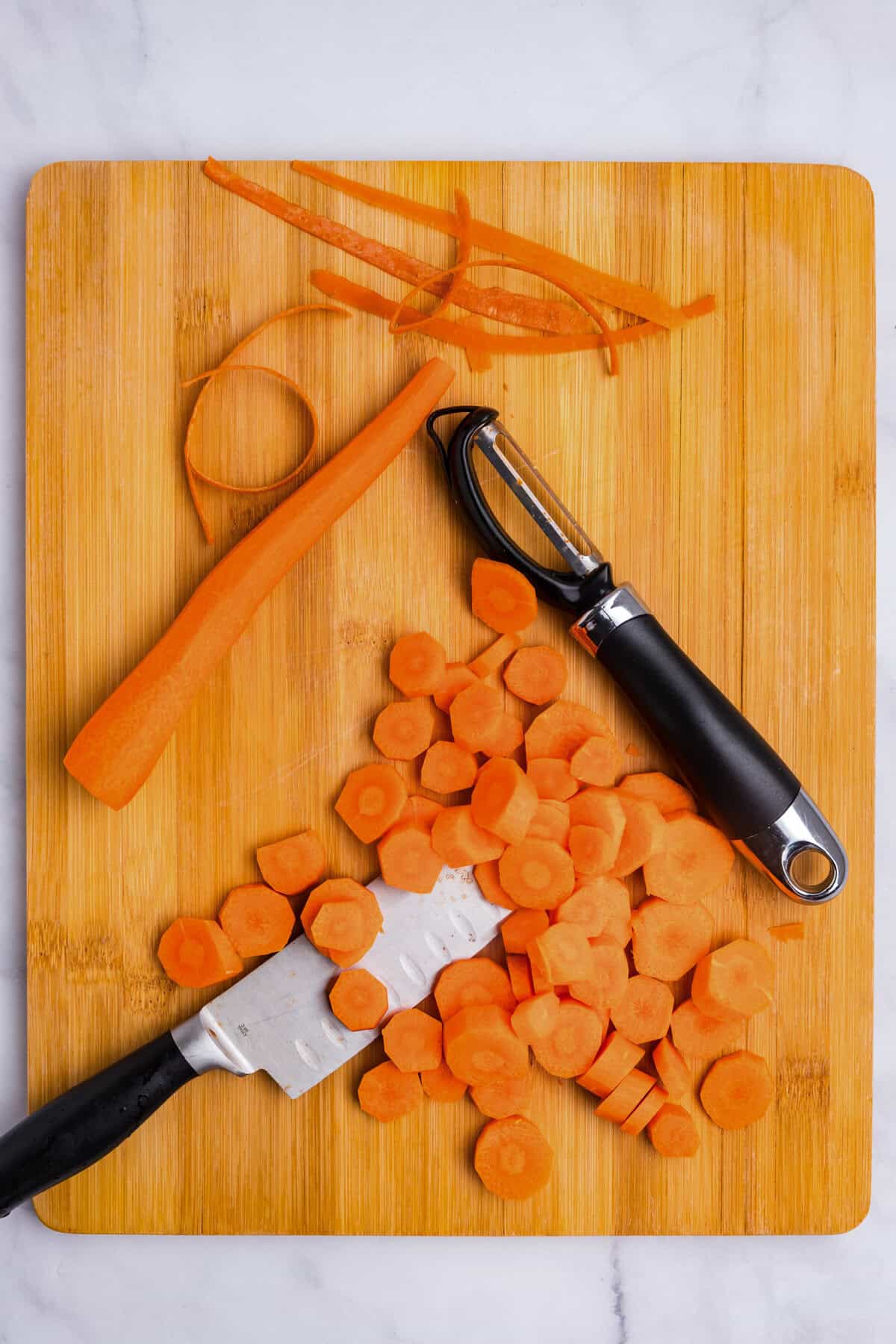Overhead picture of cut and whole carrots on a wooden cutting board with a peeler and knife positioned next to them.
