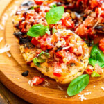 A close-up of bruschetta chicken served on a wooden cutting board. The grilled chicken breast is topped with a fresh mixture of diced tomatoes, garlic, basil, and Parmesan cheese, drizzled with balsamic glaze. Garnished with whole basil leaves and shredded Parmesan.