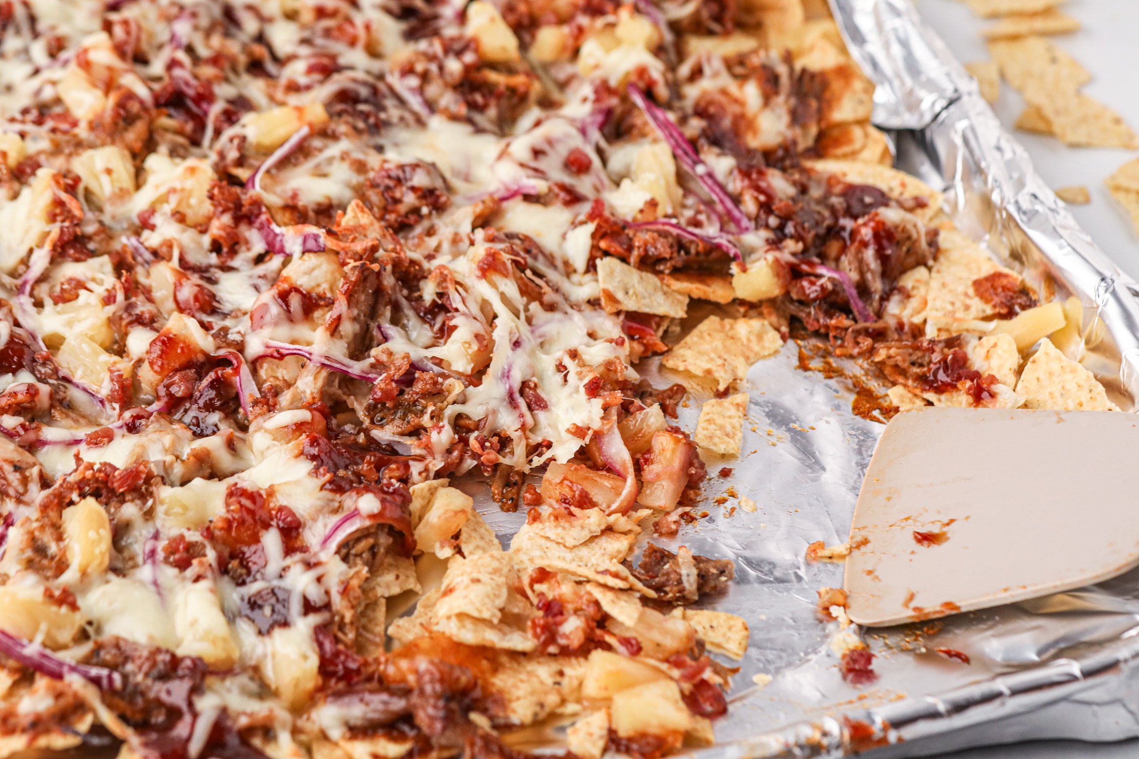 A close-up of a foil-lined baking sheet with freshly baked pulled pork nachos, topped with melted cheese, barbecue sauce, red onion slices, pineapple chunks, and crumbled bacon. A spatula rests on the sheet, and some chips are broken or scattered.