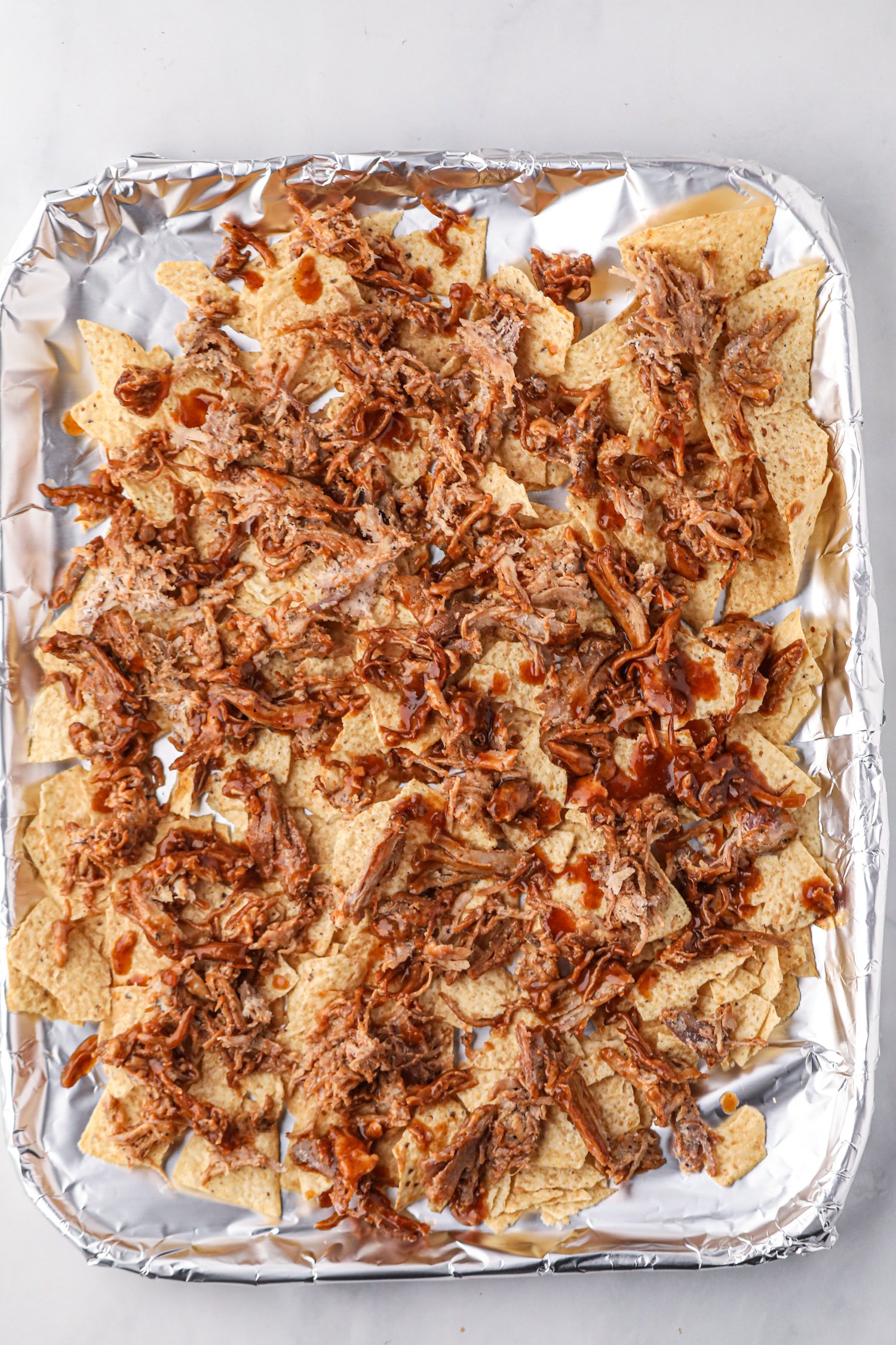 A foil-lined baking sheet filled with tortilla chips topped with shredded pulled pork and drizzled with barbecue sauce. The nachos are prepared but not yet fully assembled or baked