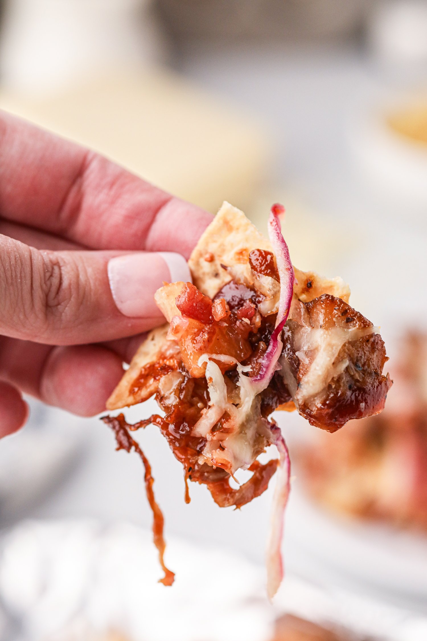 A close-up of a hand holding a loaded nacho chip topped with melted cheese, pulled pork, barbecue sauce, red onion, bacon, and pineapple. The toppings are dripping slightly, emphasizing the gooey and flavorful texture.