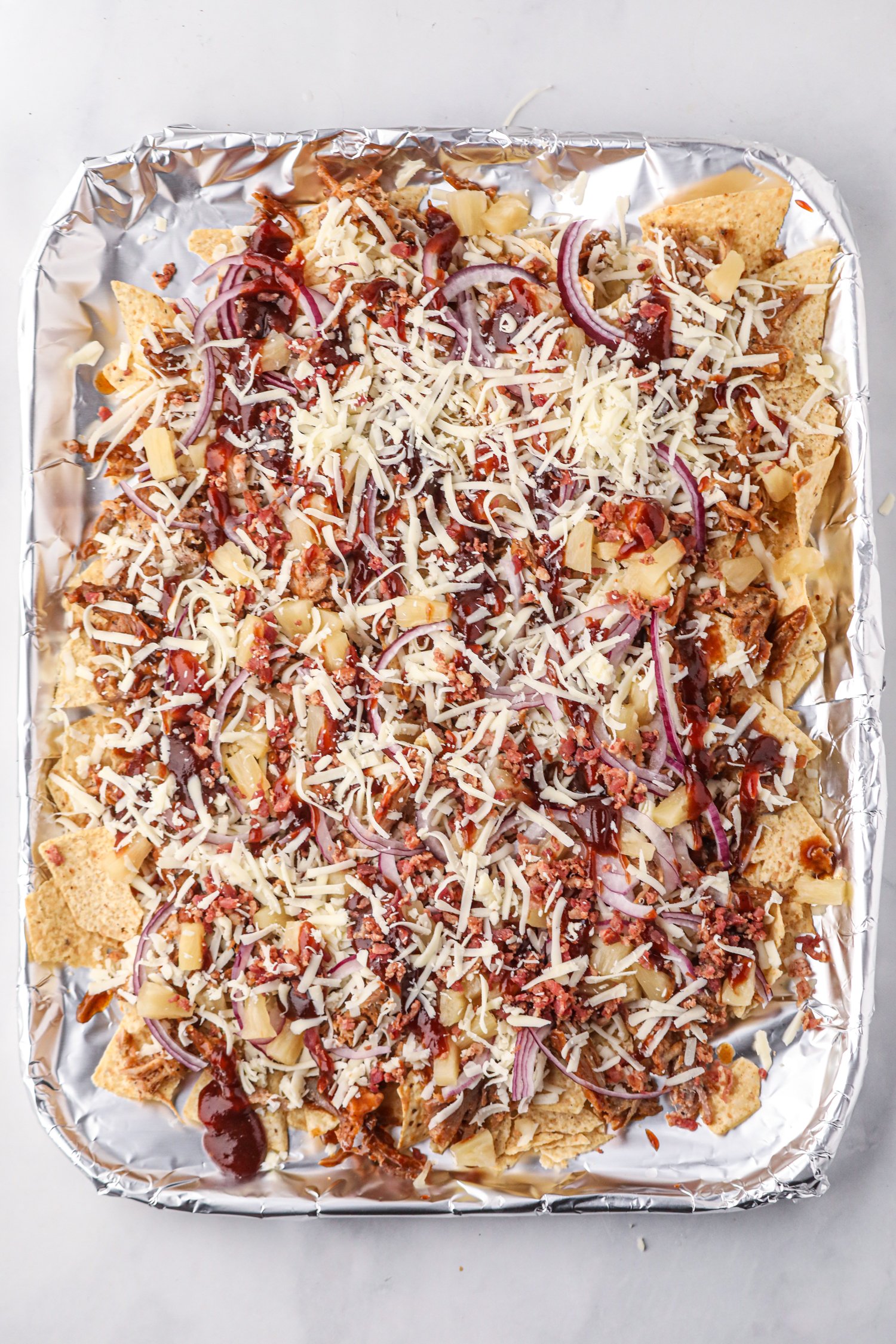 A foil-lined baking sheet filled with tortilla chips topped with shredded pulled pork, barbecue sauce, red onion slices, pineapple chunks, crumbled bacon, and shredded cheese, ready to be baked.