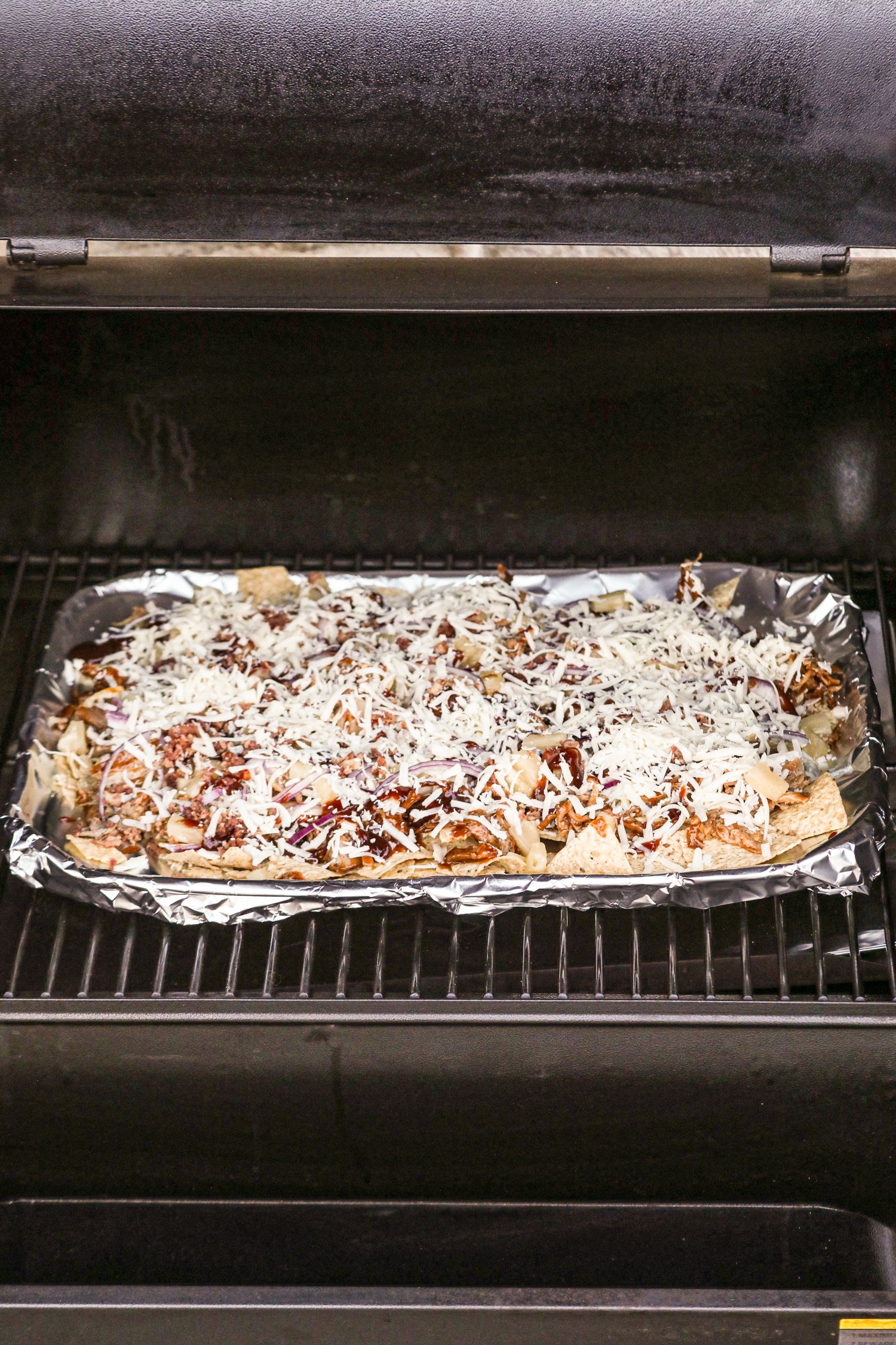 Tray of pulled pork nachos on a foil-lined baking sheet inside a smoker or grill. The nachos are topped with shredded cheese, barbecue sauce, red onions, and pulled pork, ready to be cooked. The smoker lid is partially open, showing the grill grates and interior.