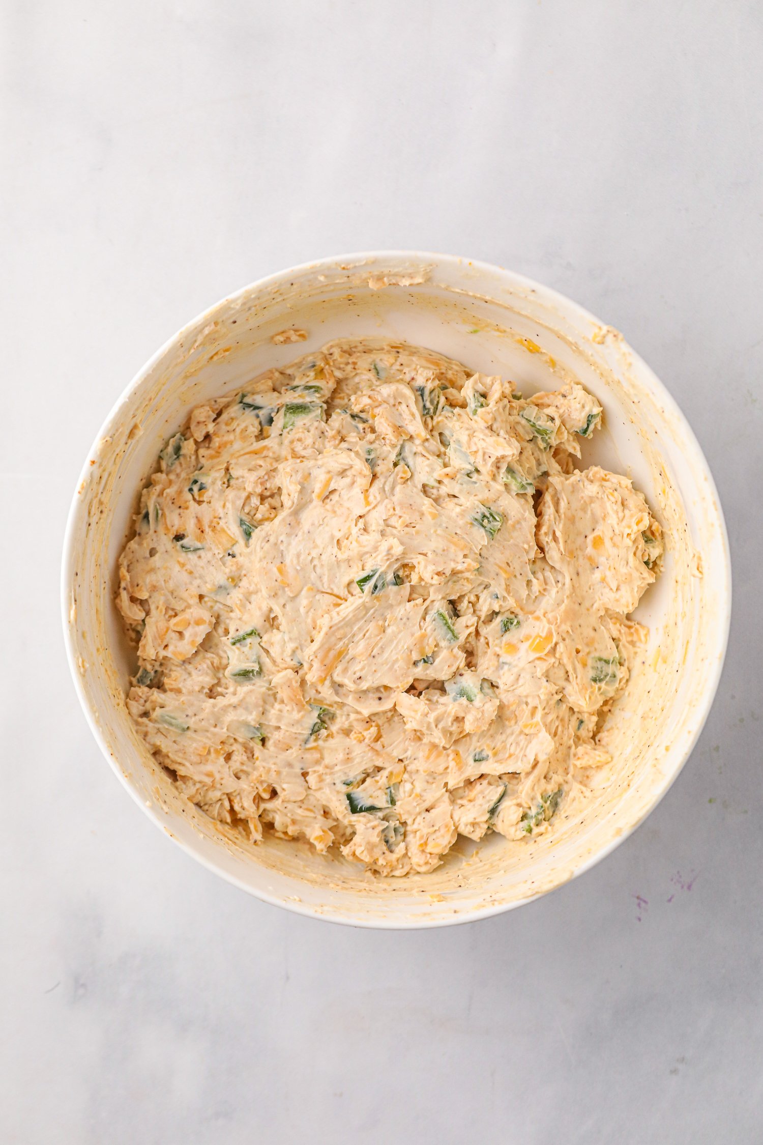 A large white mixing bowl filled with a creamy, seasoned mixture containing shredded cheese, diced green jalapeños, and spices, ready to be used for a recipe.
