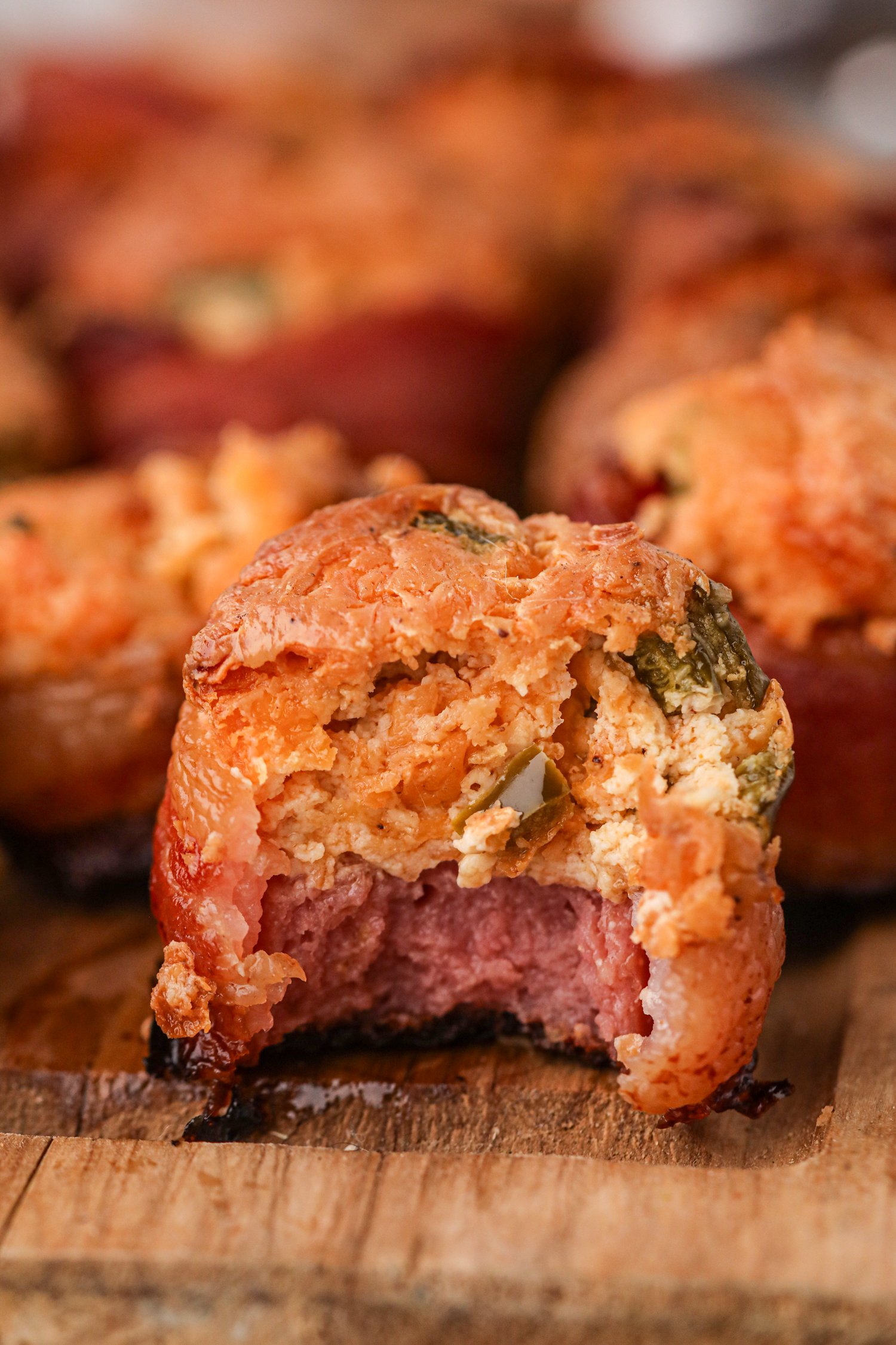 Close-up of a smoked pig shot with a bite taken out, showing its layers of crispy bacon, creamy cheese filling with diced jalapeños, and a sausage base. The filling is golden and slightly crispy on top, with flecks of seasoning visible. The background is blurred, with more pig shots on a wooden cutting board.