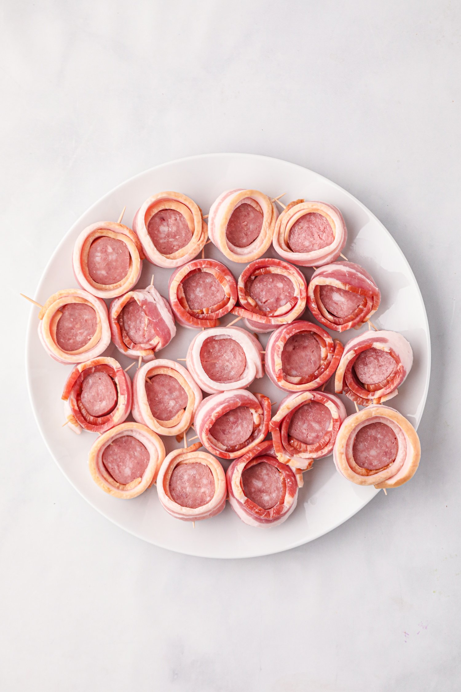 A white plate filled with uncooked smoked pig shots, which are sausage rounds wrapped in bacon and secured with toothpicks, creating small cup-like shapes ready to be stuffed and cooked.