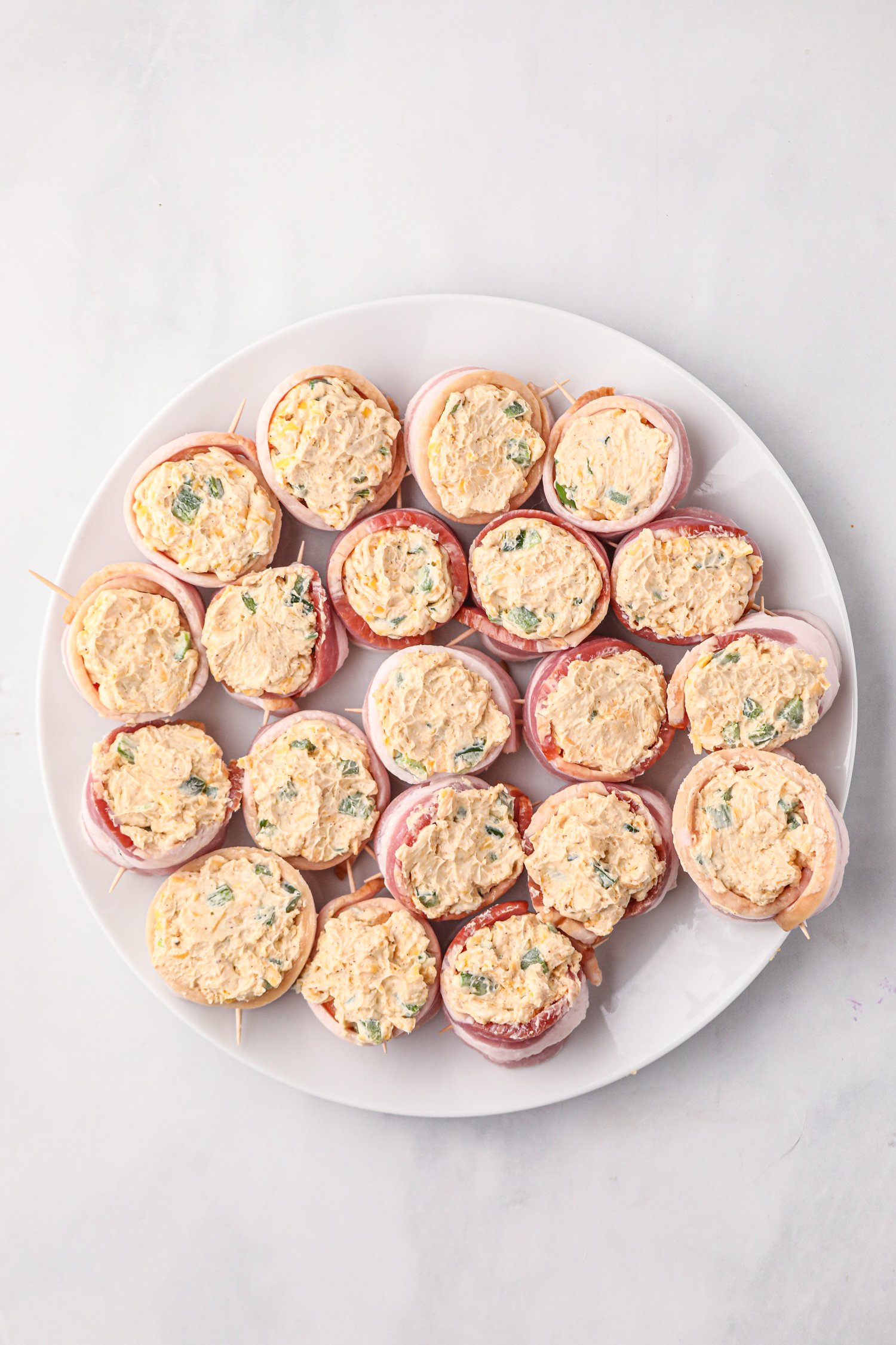 A white plate filled with uncooked smoked pig shots, which are sausage rounds wrapped in bacon and secured with toothpicks. Each bacon cup is generously stuffed with a creamy cheese mixture containing chopped green onions and seasoning, ready to be cooked.