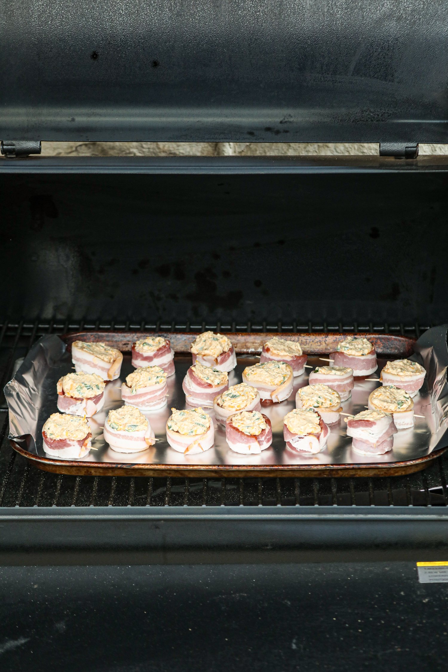 A tray of uncooked smoked pig shots placed on a foil-lined baking sheet inside a smoker. The bacon-wrapped sausage rounds are filled with a creamy cheese mixture, ready to be smoked to perfection. The smoker lid is open, showing the setup.